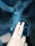 A hand with a decorative ring holds a lit cigarette between the middle and index fingers. Smoke swirls around in the air, creating a hazy atmosphere. The background is dark, emphasizing the smoke and the hand.