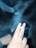 A hand with a decorative ring holds a lit cigarette between the middle and index fingers. Smoke swirls around in the air, creating a hazy atmosphere. The background is dark, emphasizing the smoke and the hand.