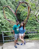 A couple stands in front of a large heart-shaped structure made of intertwining colorful branches. Behind them is lush greenery, suggesting a forest or garden setting. A sign next to them reads 'The Color of Love.' Both individuals are casually dressed, with the man in a green shirt and shorts, and the woman in a blue top and white shorts.