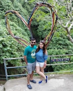 A couple stands in front of a large heart-shaped structure made of intertwining colorful branches. Behind them is lush greenery, suggesting a forest or garden setting. A sign next to them reads 'The Color of Love.' Both individuals are casually dressed, with the man in a green shirt and shorts, and the woman in a blue top and white shorts.
