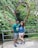 A couple stands in front of a large heart-shaped structure made of intertwining colorful branches. Behind them is lush greenery, suggesting a forest or garden setting. A sign next to them reads 'The Color of Love.' Both individuals are casually dressed, with the man in a green shirt and shorts, and the woman in a blue top and white shorts.
