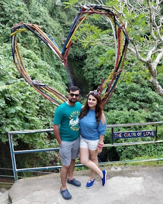 A couple stands in front of a large heart-shaped structure made of intertwining colorful branches. Behind them is lush greenery, suggesting a forest or garden setting. A sign next to them reads 'The Color of Love.' Both individuals are casually dressed, with the man in a green shirt and shorts, and the woman in a blue top and white shorts.