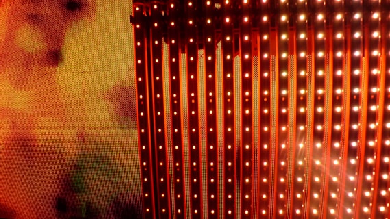 A vibrant display of vertical LED light strips emitting orange and red hues with a warm, glowing effect. The light patterns create a dynamic and colorful visual, resembling a digital or electronic art installation.