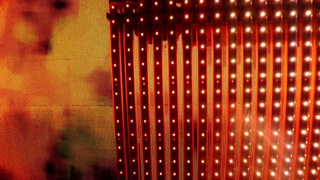 A vibrant display of vertical LED light strips emitting orange and red hues with a warm, glowing effect. The light patterns create a dynamic and colorful visual, resembling a digital or electronic art installation.