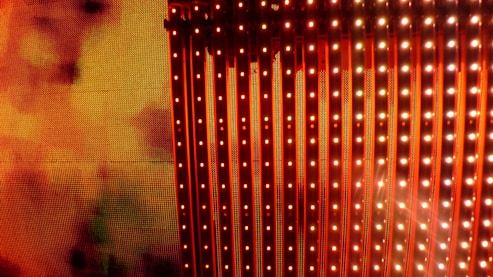 A vibrant display of vertical LED light strips emitting orange and red hues with a warm, glowing effect. The light patterns create a dynamic and colorful visual, resembling a digital or electronic art installation.