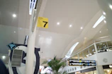 A modern airport interior with high ceilings and perforated panels. There are directional signs, including a yellow sign for Gate 7. The space is well-lit with overhead lights, and features glass railings and plants. A Wi-Fi symbol is visible, indicating connectivity services.