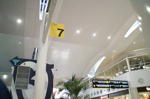 A modern airport interior with high ceilings and perforated panels. There are directional signs, including a yellow sign for Gate 7. The space is well-lit with overhead lights, and features glass railings and plants. A Wi-Fi symbol is visible, indicating connectivity services.