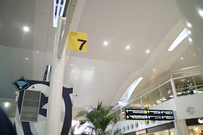 A modern airport interior with high ceilings and perforated panels. There are directional signs, including a yellow sign for Gate 7. The space is well-lit with overhead lights, and features glass railings and plants. A Wi-Fi symbol is visible, indicating connectivity services.