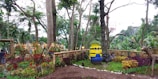 A garden area with lush greenery and various tropical plants. In the center, there is a large figure resembling a cartoon character made from stacked barrels painted yellow and blue, with a cheerful face. Surrounding this figure are colorful plants and decorative bamboo fencing. Trees rise tall in the background, with a mix of green foliage and some colorful hanging elements.