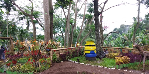 A garden area with lush greenery and various tropical plants. In the center, there is a large figure resembling a cartoon character made from stacked barrels painted yellow and blue, with a cheerful face. Surrounding this figure are colorful plants and decorative bamboo fencing. Trees rise tall in the background, with a mix of green foliage and some colorful hanging elements.