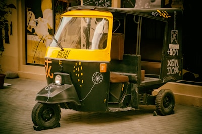 A vibrant auto rickshaw with a black and yellow color scheme is parked on a tiled street. It features artistic designs and lettering on its exterior, creating a stylish and modern look. The background includes wall art and some abstract figures, adding to the urban artistic theme.