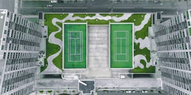 An aerial view of a rooftop featuring two green tennis courts surrounded by abstract patches of grass amidst a grey concrete surface. High-rise buildings flank the rooftop on either side, creating a symmetrical urban landscape.