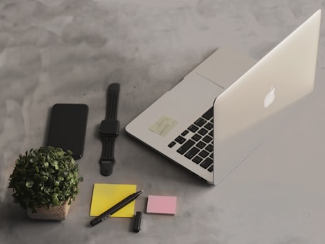 A neat and organized workspace featuring a laptop, a smartwatch, a smartphone, three sticky notes in different colors, a pen, and a small potted plant. The background is a minimalist, gray surface.