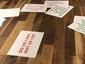 Several paper sheets are scattered on a wooden surface. The most prominent sheet displays the text 'DO AS I SAY, NOT AS I DO' in red capital letters. Other sheets contain different texts in green, partially visible.
