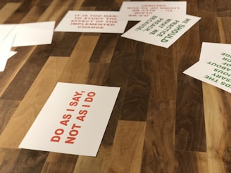 Several paper sheets are scattered on a wooden surface. The most prominent sheet displays the text 'DO AS I SAY, NOT AS I DO' in red capital letters. Other sheets contain different texts in green, partially visible.