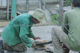 Two men are engaged in woodworking. One man wearing a green jacket and a straw hat is cutting or shaping wood while kneeling. Another man sits nearby observing or assisting. The setting appears to be outdoors, with wooden structures and greenery in the background.