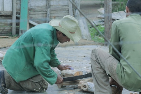 Two men are engaged in woodworking. One man wearing a green jacket and a straw hat is cutting or shaping wood while kneeling. Another man sits nearby observing or assisting. The setting appears to be outdoors, with wooden structures and greenery in the background.