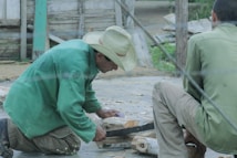 Two men are engaged in woodworking. One man wearing a green jacket and a straw hat is cutting or shaping wood while kneeling. Another man sits nearby observing or assisting. The setting appears to be outdoors, with wooden structures and greenery in the background.