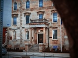 A historic stone building with an ornate facade featuring arched windows and decorative columns framing the entrance. Well-kept shrubs line the entrance steps, and several plaques are mounted on the walls. A piece of black metal chain fencing marks the sidewalk perimeter.