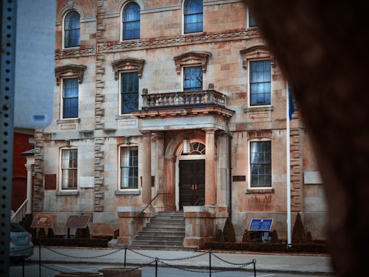 A historic stone building with an ornate facade featuring arched windows and decorative columns framing the entrance. Well-kept shrubs line the entrance steps, and several plaques are mounted on the walls. A piece of black metal chain fencing marks the sidewalk perimeter.