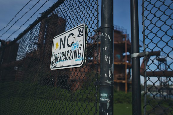 A chain-link fence with barbed wire features prominently, displaying a weathered 'No Trespassing' sign with various stickers. In the background, an industrial structure with rusted metal components is partially visible against a clear blue sky.
