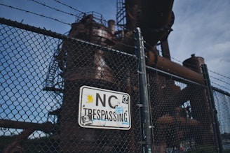 A chain-link fence with barbed wire at the top surrounds an industrial structure. A 'No Trespassing' sign is affixed to the fence, adorned with various stickers and graffiti. The industrial structure in the background appears rusted and abandoned, under a cloudy sky.