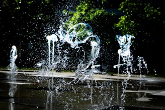 Water splashes rise from a series of fountains with droplets frozen in mid-air. The background reveals greenery with trees and a darkened area, creating a contrast with the bright, glistening water in the foreground.