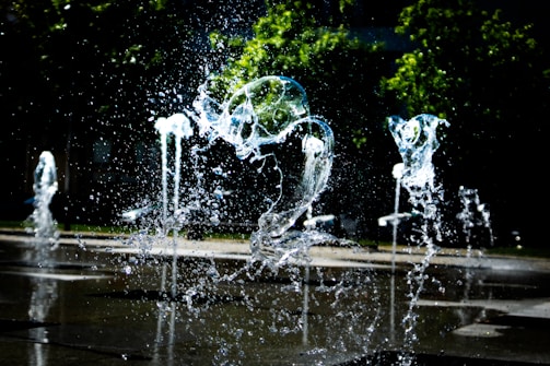 Water splashes rise from a series of fountains with droplets frozen in mid-air. The background reveals greenery with trees and a darkened area, creating a contrast with the bright, glistening water in the foreground.