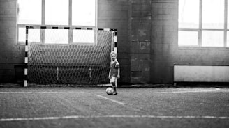 A child wearing a sports uniform stands on an indoor football field, one foot on a soccer ball. The background features a goalpost and large windows, allowing natural light to fill the space. The image is in black and white, which gives it a nostalgic and timeless feel.