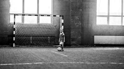 A child wearing a sports uniform stands on an indoor football field, one foot on a soccer ball. The background features a goalpost and large windows, allowing natural light to fill the space. The image is in black and white, which gives it a nostalgic and timeless feel.