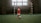 A young boy in a red soccer uniform is standing with a soccer ball on an indoor field. He is positioned in front of a goal with a net and red-and-white striped posts. The room is dimly lit with natural light coming from a window behind the goal.
