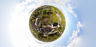 A spherical panorama provides a bird's-eye view of a small rural area, with houses, greenery, and patches of land. The landscape is surrounded by a circular horizon, blending into a bright, cloudy sky.