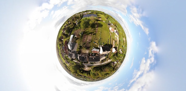 A spherical panorama provides a bird's-eye view of a small rural area, with houses, greenery, and patches of land. The landscape is surrounded by a circular horizon, blending into a bright, cloudy sky.