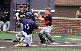 A baseball player in a purple and white uniform slides into home plate as a catcher in black and red gear attempts to tag him. Two other players and an umpire can be seen in the background, observing the play. The scene takes place on a baseball field with green and brown turf, and a brick wall behind the catcher's area.