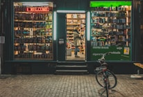 A small storefront at night displays a variety of products. The left window showcases a collection of snacks with colorful packaging under a red neon 'Welcome' sign. The center doorway is open, revealing shelves filled with bottles and boxes. A person stands inside, looking at the products. The right window also displays various items under a green neon 'Welcome Open' sign. A bicycle is parked outside on the brick pavement.