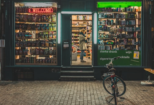 A small storefront at night displays a variety of products. The left window showcases a collection of snacks with colorful packaging under a red neon 'Welcome' sign. The center doorway is open, revealing shelves filled with bottles and boxes. A person stands inside, looking at the products. The right window also displays various items under a green neon 'Welcome Open' sign. A bicycle is parked outside on the brick pavement.