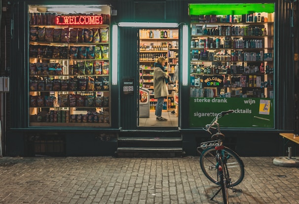 A small storefront at night displays a variety of products. The left window showcases a collection of snacks with colorful packaging under a red neon 'Welcome' sign. The center doorway is open, revealing shelves filled with bottles and boxes. A person stands inside, looking at the products. The right window also displays various items under a green neon 'Welcome Open' sign. A bicycle is parked outside on the brick pavement.
