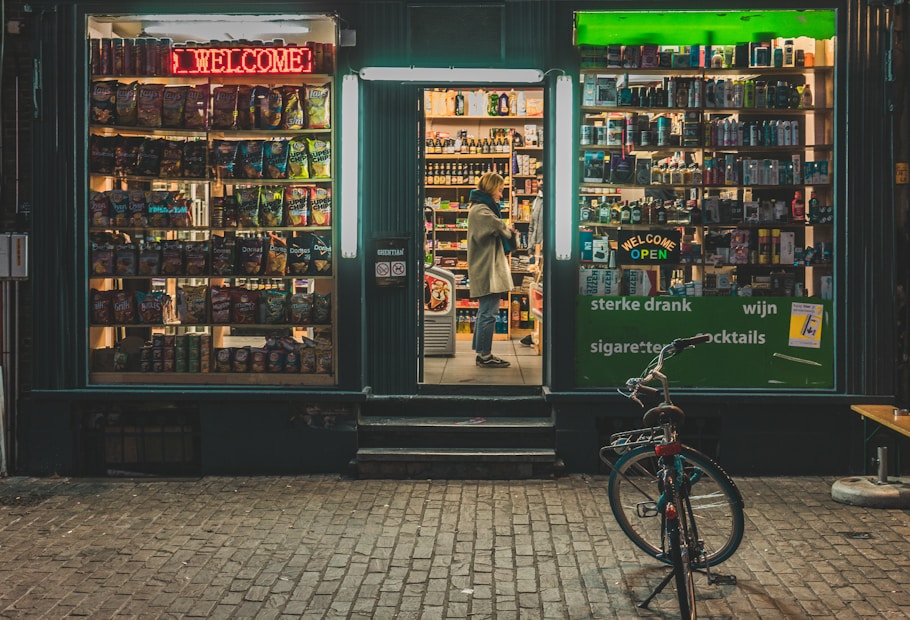 A small storefront at night displays a variety of products. The left window showcases a collection of snacks with colorful packaging under a red neon 'Welcome' sign. The center doorway is open, revealing shelves filled with bottles and boxes. A person stands inside, looking at the products. The right window also displays various items under a green neon 'Welcome Open' sign. A bicycle is parked outside on the brick pavement.