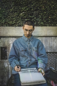A person wearing glasses and a denim shirt sits on a bench, intently sketching with a pencil in an open notebook. The background features a textured stone wall partially covered with green foliage.