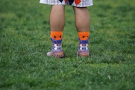 A person's legs are visible from the knees down, standing on a grassy surface. They are wearing colorful socks with orange, blue, and purple patterns and athletic shoes. The person is dressed in sportswear, suggesting an athletic or recreational setting.