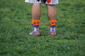 A person's legs are visible from the knees down, standing on a grassy surface. They are wearing colorful socks with orange, blue, and purple patterns and athletic shoes. The person is dressed in sportswear, suggesting an athletic or recreational setting.