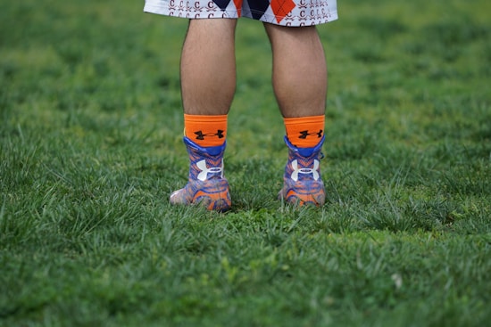 A person's legs are visible from the knees down, standing on a grassy surface. They are wearing colorful socks with orange, blue, and purple patterns and athletic shoes. The person is dressed in sportswear, suggesting an athletic or recreational setting.