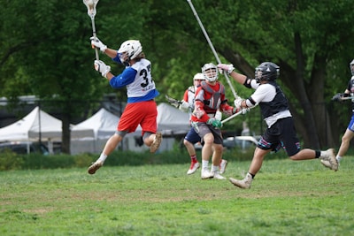 Several lacrosse players are actively engaged in a game on a grass field, surrounded by trees and tents in the background. One player in red shorts is jumping mid-air holding a lacrosse stick, while other players in protective gear and helmets are also involved in the action.