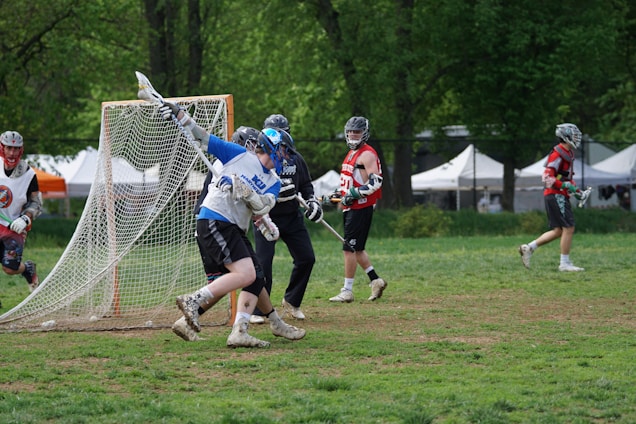 A group of lacrosse players is engaged in a game on a grassy field surrounded by trees. One player is in motion near the goal, holding a lacrosse stick, while others are stationed around the goal area. They are wearing protective helmets, gloves, and jerseys with various colors. Tents are seen in the background, suggesting an event or tournament setting.