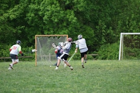Four lacrosse players are on a field with two goals visible. Three players are wearing helmets and protective gear while actively engaged in play. One player appears to be attempting a goal shot.