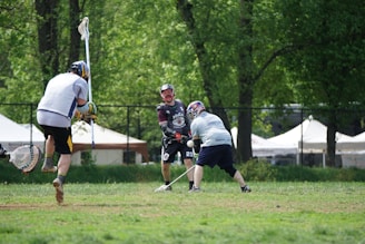 Three men are playing lacrosse on a grassy field with a backdrop of trees and white tents. They are wearing protective gear, including helmets and gloves. One player in a dark jersey faces the others and is focused on the ball in motion.