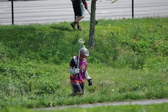 A person wearing a helmet and protective gear carries a long stick while walking on lush green grass. Another person in the background trims the grass with a string trimmer near a chain-link fence.