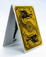 A playing card is displayed in an upright, tent-like position. The card features a golden, intricate design of a dragon on a black background, surrounded by ornate patterns. The style is reminiscent of traditional Asian art.