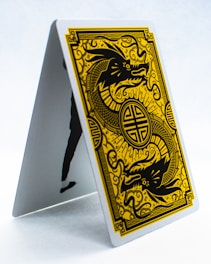 A playing card is displayed in an upright, tent-like position. The card features a golden, intricate design of a dragon on a black background, surrounded by ornate patterns. The style is reminiscent of traditional Asian art.