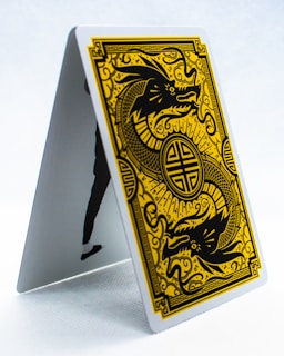 A playing card is displayed in an upright, tent-like position. The card features a golden, intricate design of a dragon on a black background, surrounded by ornate patterns. The style is reminiscent of traditional Asian art.